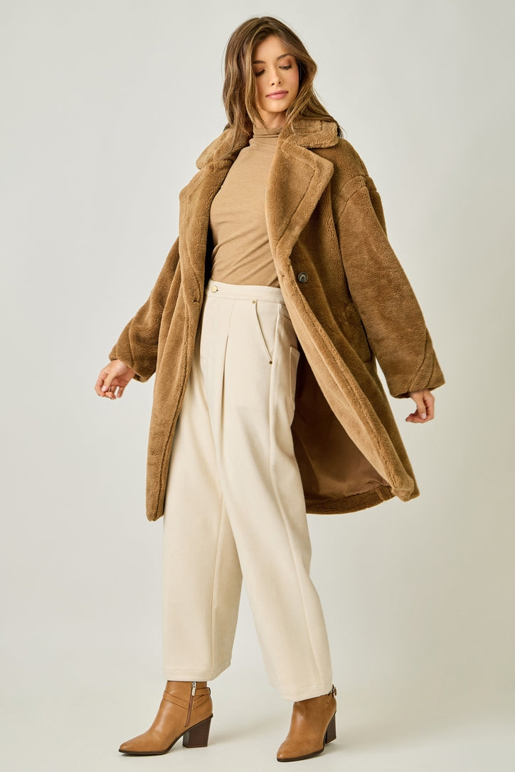 Woman wearing a brown coat, beige sweater, and white pants on a plain background